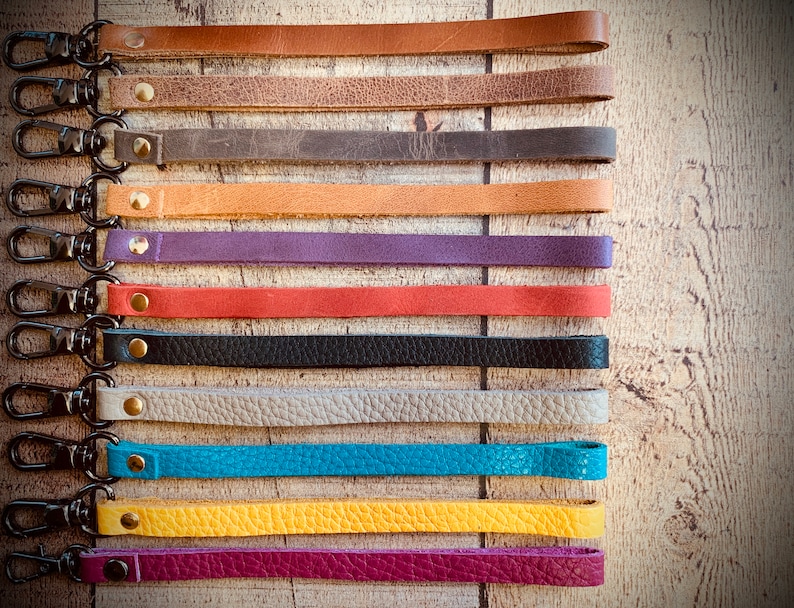 Op de afbeelding: Een verzameling leren polsbandjes in diverse kleuren, waaronder bruin, grijs, oranje, paars, rood, zwart, beige, turquoise, geel en magenta. Elke band heeft een metalen sluiting en een messing klinknagel.