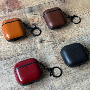 May include: Four protective cases for wireless earbuds, each featuring a smooth finish in tan, dark brown, deep red, or black. Each case includes a black metal hinge, a small circular cutout for a charging port, and a black metal keyring.