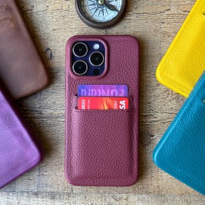 Peut inclure: Une coque de t&eacute;l&eacute;phone en cuir textur&eacute; bordeaux avec un porte-cartes contenant deux cartes bancaires. La coque pr&eacute;sente une d&eacute;coupe pour triple appareil photo. Autour, d'autres coques en cuir marron, jaune, violet et bleu sarcelle sont visibles.