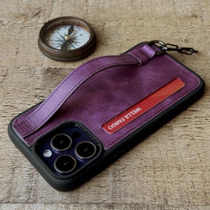 May include: A purple leather phone case with a strap and a Wells Fargo card. The case has a black border and a chain attachment. A vintage compass sits in the background on a wooden surface.