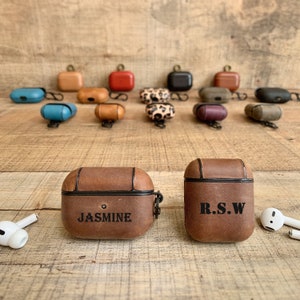 May include: Two brown leather Airpod cases with personalised names. The case on the left says "JASMINE" and the case on the right says "R.S.W."
