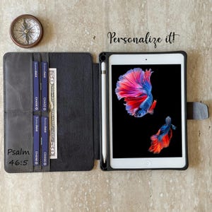 May include: A black iPad case with a stylus, credit cards, and a fifty-dollar bill. The iPad screen shows two vibrant betta fish on a black backdrop. The text "Personalize it!" is above the iPad, and "Psalm 46:5" is on the case.
