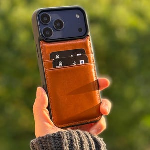 Peut inclure: Une coque de smartphone noire avec un porte-cartes en cuir marron clair fix&eacute; au dos. Le porte-cartes contient deux cartes bancaires noires avec du texte blanc visible. La coque pr&eacute;sente une d&eacute;coupe pour le triple capteur photo et le flash.