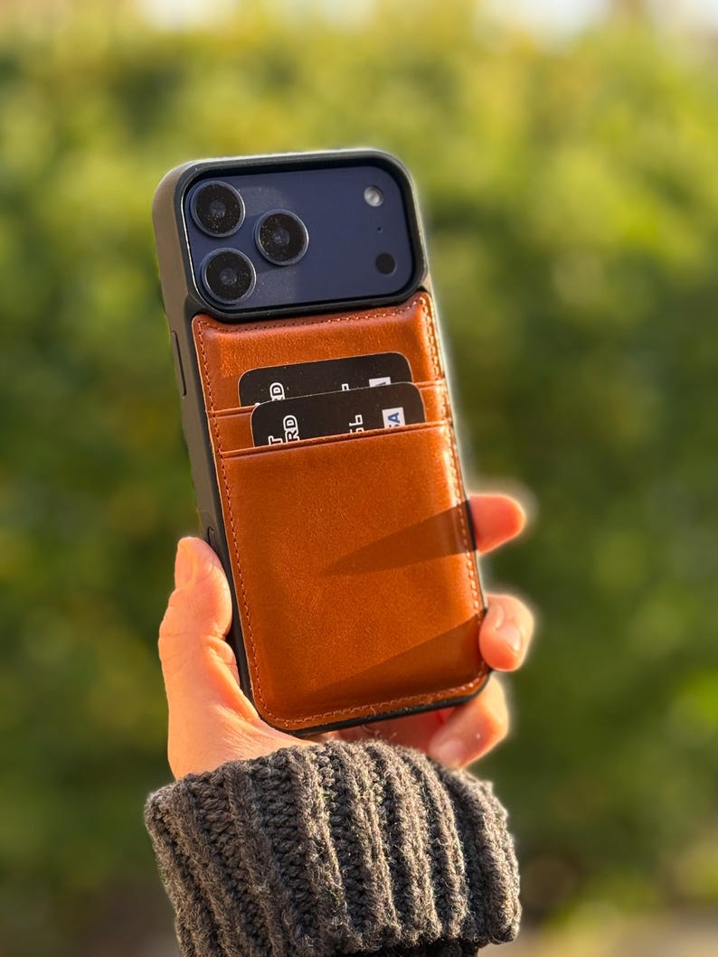 Peut inclure: Une coque de smartphone noire avec un porte-cartes en cuir marron clair fix&eacute; au dos. Le porte-cartes contient deux cartes bancaires noires avec du texte blanc visible. La coque pr&eacute;sente une d&eacute;coupe pour le triple capteur photo et le flash.