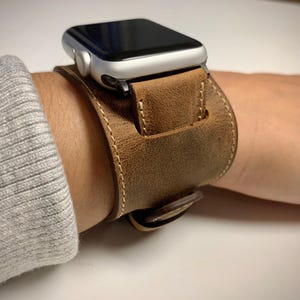 Peut inclure: Une montre connect&eacute;e argent&eacute;e avec un &eacute;cran noir fix&eacute;e sur un large bracelet manchette en cuir marron. Le bracelet en cuir pr&eacute;sente des coutures claires visibles sur les bords et une fermeture &agrave; boucle s&eacute;curis&eacute;e autour du poignet.