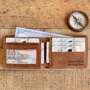 May include: Open brown leather wallet with a $100 bill, a driver's license, and several cards. The wallet has the text "Engrave Your Message Here" and is next to a compass on a wooden surface.