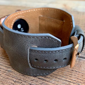 Peut inclure: Un bracelet de montre large en cuir gris textur&eacute; avec une boucle en m&eacute;tal dor&eacute; rose et une doublure int&eacute;rieure en cuir beige. Le bracelet est con&ccedil;u pour entourer une montre connect&eacute;e, r&eacute;v&eacute;lant le capteur circulaire noir &agrave; l'int&eacute;rieur.