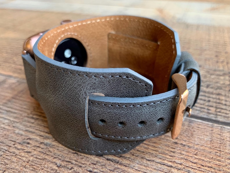 Peut inclure: Un bracelet de montre large en cuir gris textur&eacute; avec une boucle en m&eacute;tal dor&eacute; rose et une doublure int&eacute;rieure en cuir beige. Le bracelet est con&ccedil;u pour entourer une montre connect&eacute;e, r&eacute;v&eacute;lant le capteur circulaire noir &agrave; l'int&eacute;rieur.