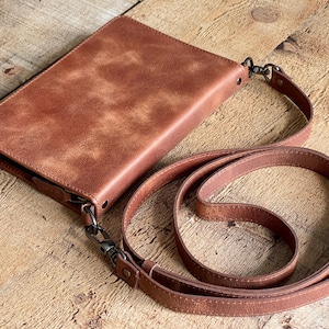 May include: A rectangular brown leather crossbody bag with a smooth, mottled finish. The bag features a top zipper closure, dark metal hardware, and a matching adjustable leather shoulder strap attached with swivel clips on both sides.