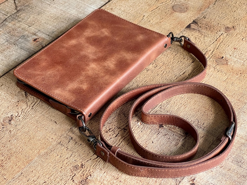 May include: A rectangular brown leather crossbody bag with a smooth, mottled finish. The bag features a top zipper closure, dark metal hardware, and a matching adjustable leather shoulder strap attached with swivel clips on both sides.