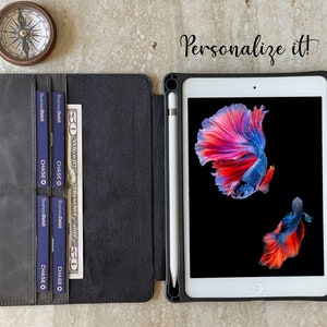 May include: A black leather iPad case with a white stylus, credit cards, and a fifty-dollar bill. The iPad displays a colorful betta fish image. A compass is in the upper left corner. Text reads "Personalize it!"