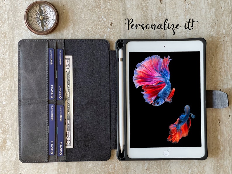 May include: A black leather iPad case with a white stylus, credit cards, and a fifty-dollar bill. The iPad displays a colorful betta fish image. A compass is in the upper left corner. Text reads "Personalize it!"