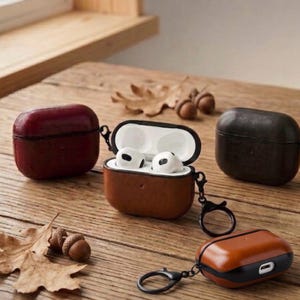 Pode incluir: Uma variedade de capas de couro para AirPods em várias cores, incluindo castanho, vermelho escuro e preto. Uma capa está aberta, revelando fones de ouvido brancos. As capas têm uma aparência texturizada e são exibidas em uma superfície de madeira.
