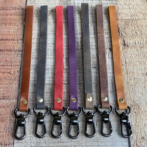 May include: Seven leather wrist straps in various colors: brown, gray, red, purple, and tan. Each strap has a black metal clasp and is secured with a brass rivet. The straps are arranged on a wooden surface.
