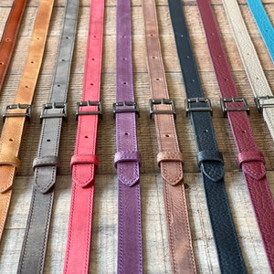 May include: A collection of ten adjustable leather straps in various colors including brown, tan, gray, red, purple, black, burgundy, beige, and turquoise. Each strap features a dark metal buckle, a sliding keeper, and pre-punched holes for sizing.
