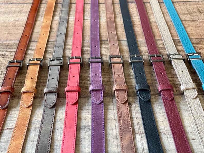 May include: A collection of ten adjustable leather straps in various colors including brown, tan, gray, red, purple, black, burgundy, beige, and turquoise. Each strap features a dark metal buckle, a sliding keeper, and pre-punched holes for sizing.