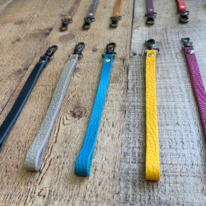 Op de afbeelding: Leren polsbandjes in diverse kleuren, waaronder zwart, beige, blauw, geel en roze. Elke band heeft een metalen sluiting om aan een tas of ander item te bevestigen. De bandjes zijn gerangschikt op een houten oppervlak.