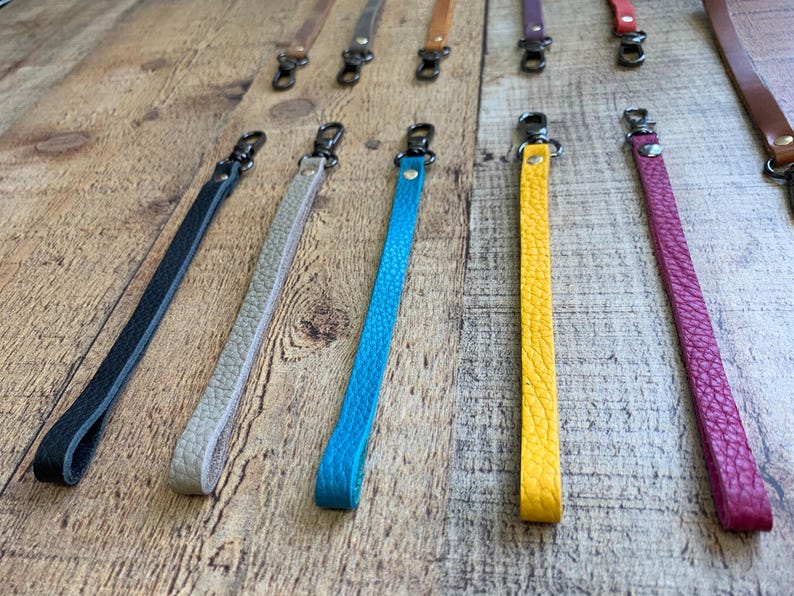 Op de afbeelding: Leren polsbandjes in diverse kleuren, waaronder zwart, beige, blauw, geel en roze. Elke band heeft een metalen sluiting om aan een tas of ander item te bevestigen. De bandjes zijn gerangschikt op een houten oppervlak.