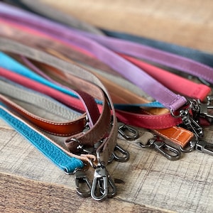 May include: A collection of leather bag straps in various colors including turquoise, brown, red, purple, and black. Each strap features a textured surface and is equipped with dark metal swivel lobster claw clasps at both ends for attachment.