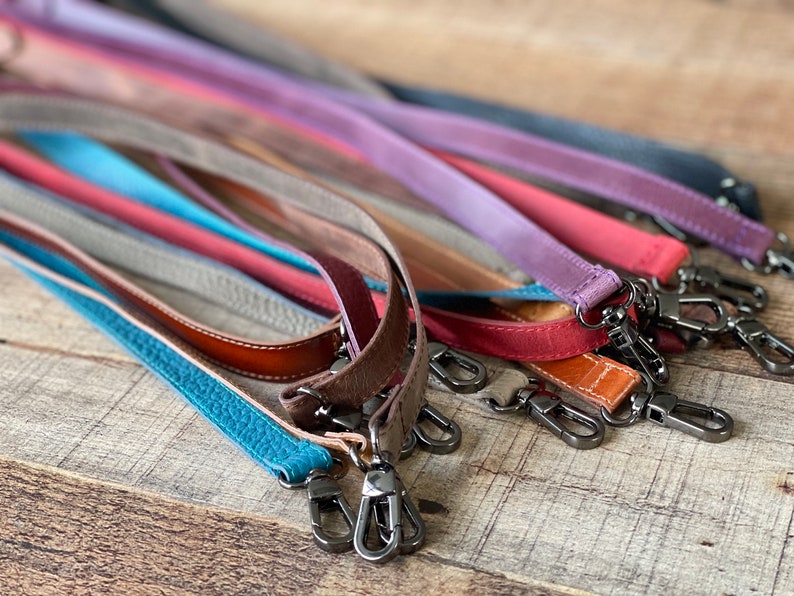 May include: A collection of leather bag straps in various colors including turquoise, brown, red, purple, and black. Each strap features a textured surface and is equipped with dark metal swivel lobster claw clasps at both ends for attachment.