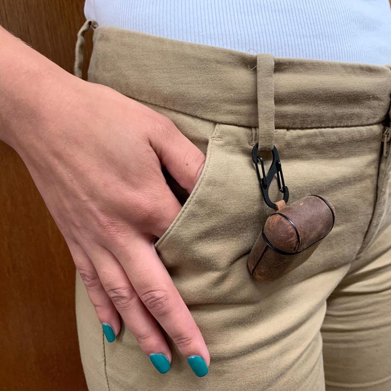 May include: A brown leather protective case for wireless earbuds featuring a black metal carabiner clip attached to a belt loop on tan trousers. The case has a smooth, rounded rectangular shape with visible stitching and a secure closure mechanism.