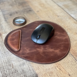 Puede incluir: Un mouse pad de cuero marrón con un borde cosido y un acento marrón más claro. Un ratón inalámbrico negro está sobre la almohadilla. Una brújula está en el fondo.