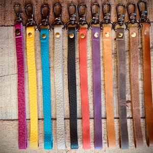 Op de afbeelding: Een verzameling leren polsbandjes in diverse kleuren, waaronder paars, geel, turquoise, wit, zwart, koraal, paars, bruin, grijs en bruin. Elk bandje heeft een metalen sluiting en een klinknagel. De bandjes liggen op een houten ondergrond.