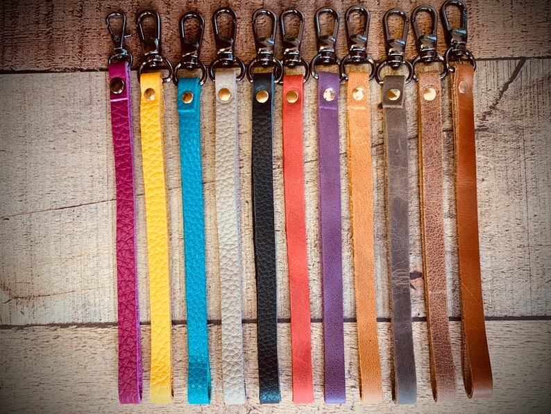 Op de afbeelding: Een verzameling leren polsbandjes in diverse kleuren, waaronder paars, geel, turquoise, wit, zwart, koraal, paars, bruin, grijs en bruin. Elk bandje heeft een metalen sluiting en een klinknagel. De bandjes liggen op een houten ondergrond.