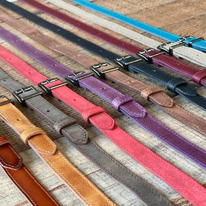 May include: A collection of narrow leather straps in various colors including brown, tan, gray, red, purple, black, burgundy, beige, and blue. Each strap features a metal buckle, a matching leather keeper loop, and pre-punched adjustment holes.