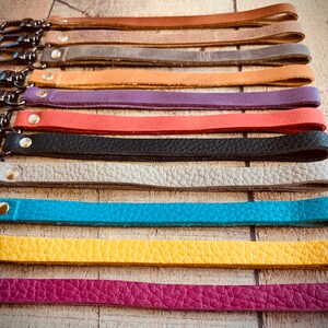 Op de afbeelding: Een verzameling kleurrijke leren polsbandjes met zwarte metalen draaisluitingen. De bandjes zijn er in verschillende kleuren, waaronder bruin, grijs, bruin, paars, rood, zwart, lichtgrijs, turquoise, geel en magenta. Elke band is vastgezet met een metalen klinknagel.