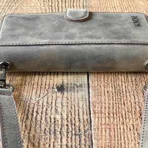 May include: A rectangular gray leather wallet with a magnetic snap closure and a detachable shoulder strap. The wallet features dark metal hardware clips on both sides and the letters KMM embossed in the upper right corner of the front surface.