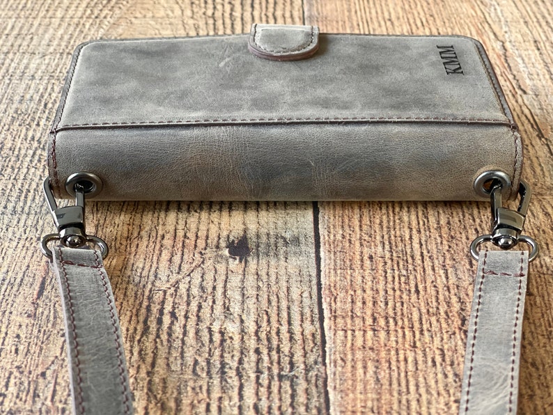 May include: A rectangular gray leather wallet with a magnetic snap closure and a detachable shoulder strap. The wallet features dark metal hardware clips on both sides and the letters KMM embossed in the upper right corner of the front surface.
