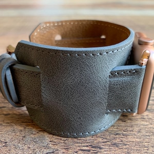 Peut inclure: Un bracelet de montre en cuir gris antique avec un design large et des coutures visibles. Le bracelet comporte un connecteur dor&eacute; m&eacute;tallique fix&eacute; &agrave; un bo&icirc;tier de montre connect&eacute;e. L'int&eacute;rieur du bracelet est beige et l'ext&eacute;rieur est textur&eacute;.
