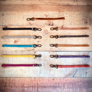 Op de afbeelding: Een verzameling leren polsbandjes in diverse kleuren, waaronder zwart, bruin, grijs, blauw, geel, paars en rood. Elk bandje heeft een metalen sluiting voor eenvoudige bevestiging. De bandjes liggen op een houten ondergrond.