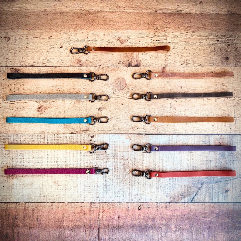 Op de afbeelding: Een verzameling leren polsbandjes in diverse kleuren, waaronder zwart, bruin, grijs, blauw, geel, paars en rood. Elk bandje heeft een metalen sluiting voor eenvoudige bevestiging. De bandjes liggen op een houten ondergrond.