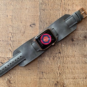 Peut inclure: Une montre connect&eacute;e avec un bo&icirc;tier dor&eacute; et un &eacute;cran num&eacute;rique affichant un motif floral g&eacute;om&eacute;trique rouge et bleu, fix&eacute;e sur un large bracelet manchette en cuir gris avec une boucle en m&eacute;tal dor&eacute; et plusieurs trous de r&eacute;glage.
