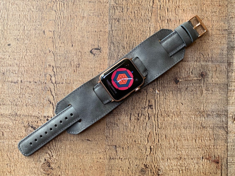 Peut inclure: Une montre connect&eacute;e avec un bo&icirc;tier dor&eacute; et un &eacute;cran num&eacute;rique affichant un motif floral g&eacute;om&eacute;trique rouge et bleu, fix&eacute;e sur un large bracelet manchette en cuir gris avec une boucle en m&eacute;tal dor&eacute; et plusieurs trous de r&eacute;glage.
