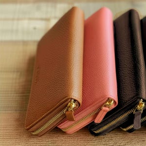 May include: A close-up of several leather wallets in various colors, including tan, coral, and dark brown. Each wallet has a gold-colored zipper closure. The wallets are arranged side by side, showcasing their different hues and textures.