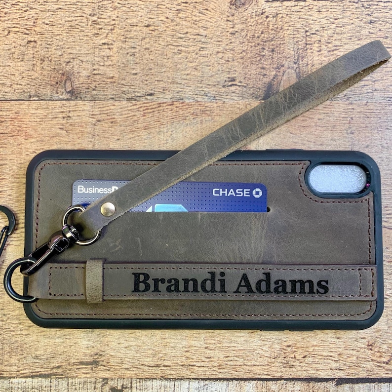 Op de afbeelding: Een bruine leren telefoonhoes met een riem en een kaartsleuf. De hoes is gegraveerd met de naam "Brandi Adams". Een creditcard is zichtbaar in de sleuf. De hoes heeft ook een polsbandje met een metalen sluiting.