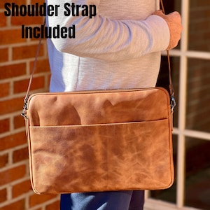 Może przedstawiać: Brązowa skórzana torba na laptopa z paskiem na ramię. Torba posiada przednią kieszeń i zapięcie na zamek błyskawiczny. Tekst "Shoulder Strap Included" jest widoczny. Zaprojektowana do przenoszenia laptopa i innych niezbędnych rzeczy.