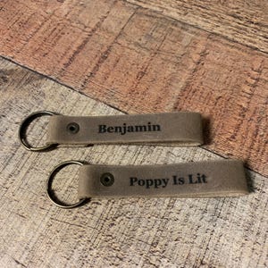May include: Two gray leather keychains with metal rings. One keychain reads "Benjamin" and the other reads "Poppy Is Lit" in black lettering. The keychains are on a wooden surface.
