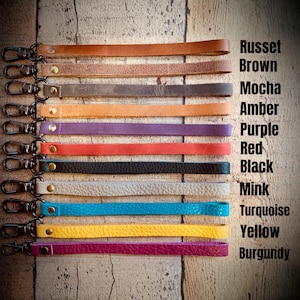 Op de afbeelding: Een verzameling leren polsbandjes in diverse kleuren, waaronder roestbruin, mokka, amber, paars, rood, zwart, nertsbruin, turquoise, geel en bordeaux. Elk bandje heeft een metalen sluiting en wordt getoond tegen een houten achtergrond. De tekst rechts vermeldt de kleurnamen.