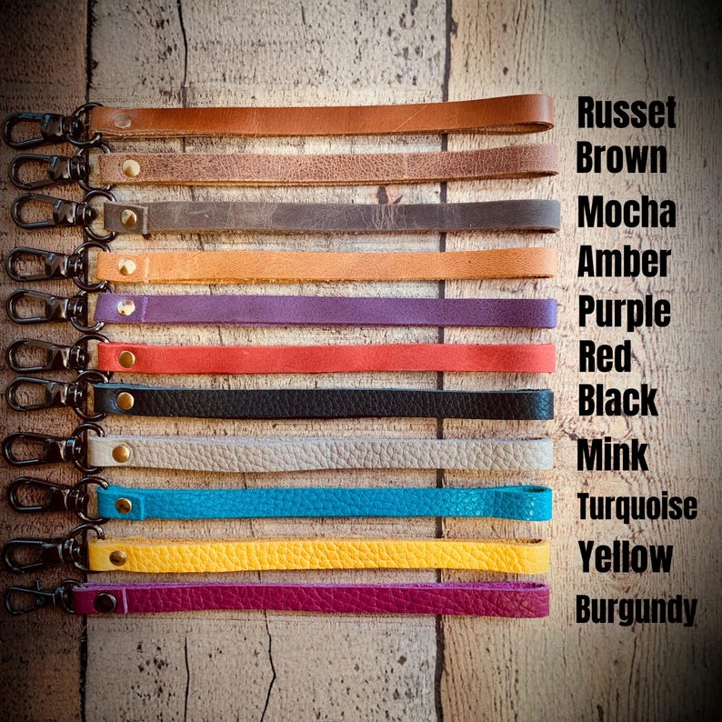 Op de afbeelding: Een verzameling leren polsbandjes in diverse kleuren, waaronder roestbruin, mokka, amber, paars, rood, zwart, nertsbruin, turquoise, geel en bordeaux. Elk bandje heeft een metalen sluiting en wordt getoond tegen een houten achtergrond. De tekst rechts vermeldt de kleurnamen.