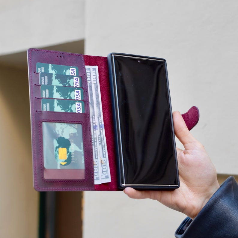 Puede incluir: Una funda de tel&eacute;fono de cuero color ciruela con dise&ntilde;o de cartera. La funda est&aacute; abierta, revelando tarjetas de cr&eacute;dito, una ventana de identificaci&oacute;n transparente y varios billetes de cien d&oacute;lares. Un tel&eacute;fono inteligente negro se sostiene en el lado derecho de la funda.