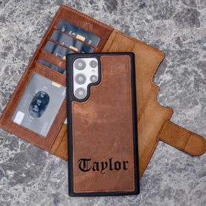Puede incluir: Funda de tel&eacute;fono de cuero marr&oacute;n con el nombre "Taylor" en fuente g&oacute;tica. La funda est&aacute; junto a una cartera de cuero marr&oacute;n con ranuras para tarjetas. La funda del tel&eacute;fono tiene un borde negro y un recorte para la c&aacute;mara.