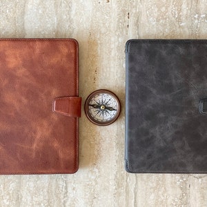 May include: Two leather tablet cases, one brown and one gray, with magnetic closures. A small compass sits between them. The cases have a textured appearance, suggesting a vintage or rustic style. The compass has a detailed face.