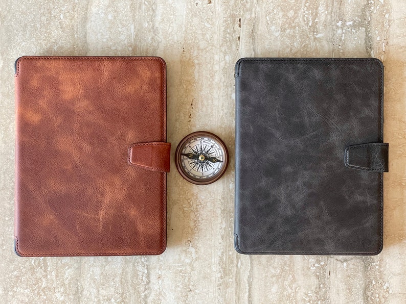 May include: Two leather tablet cases, one brown and one gray, with magnetic closures. A small compass sits between them. The cases have a textured appearance, suggesting a vintage or rustic style. The compass has a detailed face.