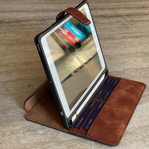 May include: A brown leather tablet case with a black frame, open to display the screen. The case has a magnetic closure and slots for cards. The tablet screen reflects a blurred image.