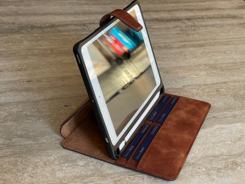 May include: A brown leather tablet case with a black frame, open to display the screen. The case has a magnetic closure and slots for cards. The tablet screen reflects a blurred image.