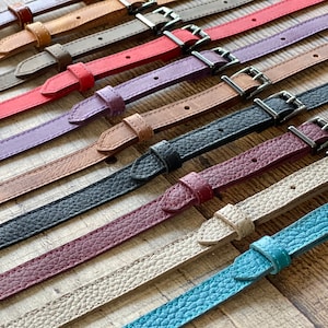 May include: A collection of thin leather straps in various colors including brown, tan, gray, red, purple, black, burgundy, beige, and teal. Each strap features a matching leather keeper, a pointed tip, and a silver-toned metal buckle.